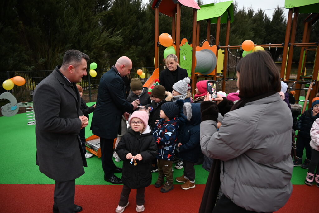 Grupa dzieci w zimowych ubraniach na placu zabaw z kolorowymi balonami, obok dwóch mężczyzn i kobiety, jedna osoba robi zdjęcie telefonem.