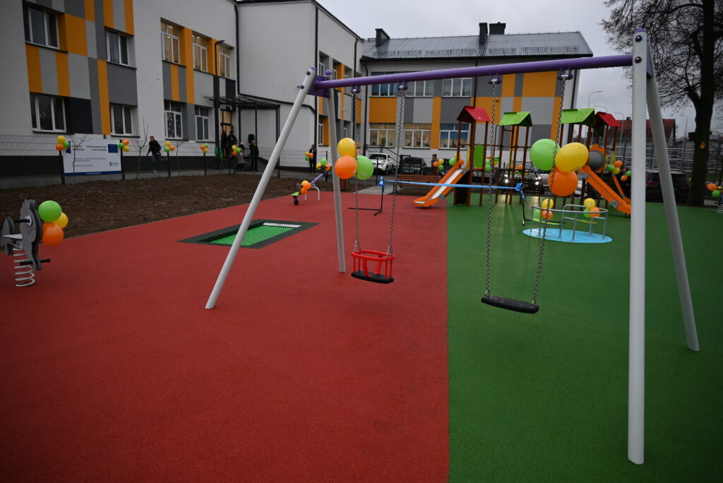 Plac zabaw z czerwoną i zieloną nawierzchnią, huśtawki ozdobione kolorowymi balonami, zjeżdżalnie, budynek z żółto-szarymi pasami w tle.