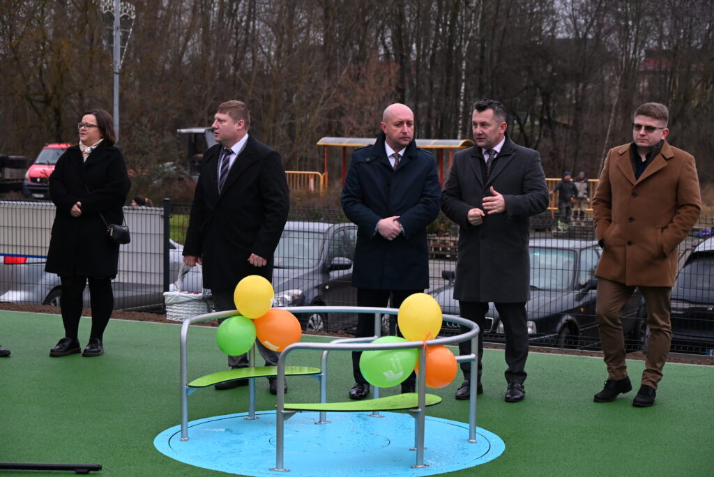 Pięć osób w ciemnych płaszczach stoi na zielonej nawierzchni obok okrągłej konstrukcji z metalowych rur z zielonymi i pomarańczowymi balonami, w tle ogrodzenie i samochody.