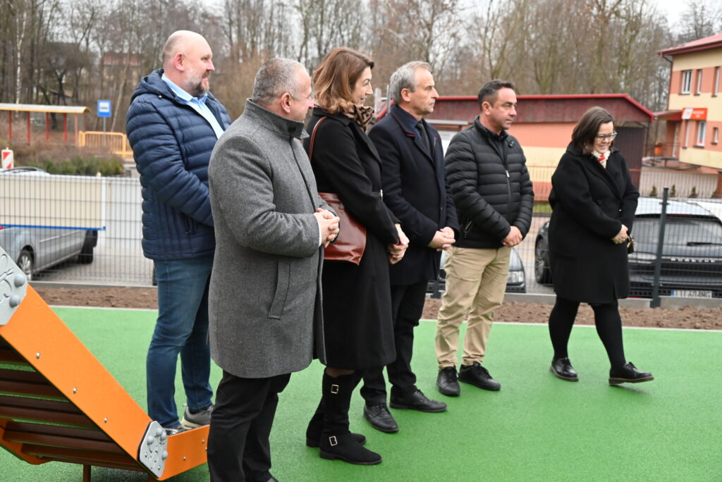 Grupa sześciu osób ubranych w płaszcze i kurtki stoi na zielonej nawierzchni na zewnątrz, w tle ogrodzenie i budynki.