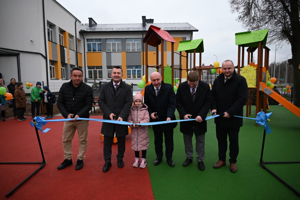 Grupa mężczyzn i dziewczynka na placu zabaw z kolorową nawierzchnią, przecinają niebieską wstęgę, w tle budynek i sprzęt placu zabaw z zielonymi i czerwonymi elementami.