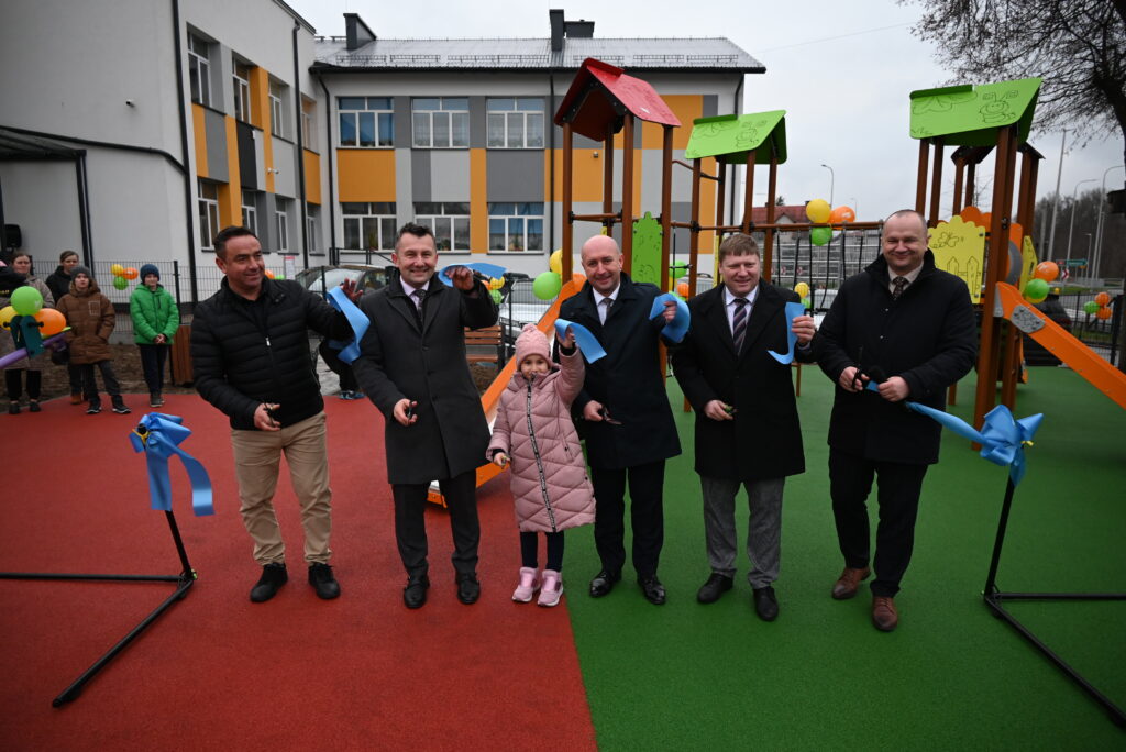 Grupa mężczyzn i dziewczynka na kolorowym placu zabaw z zieloną i czerwoną nawierzchnią, trzymający pocięte niebieskie wstążki, w tle budynek i elementy placu zabaw.