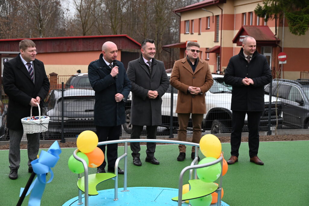 Pięciu mężczyzn w płaszczach stoi na zielonym placu zabaw obok karuzeli ozdobionej żółtymi, zielonymi i pomarańczowymi balonami, w tle budynek i samochody.
