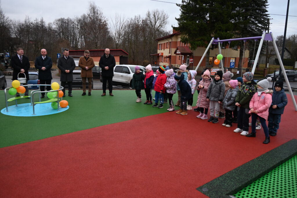 Dzieci w kolorowych kurtkach i czapkach stoją na placu zabaw z czerwoną i zieloną nawierzchnią, obok huśtawki i karuzeli ozdobionej balonami, w tle dorośli w ciemnych płaszczach.