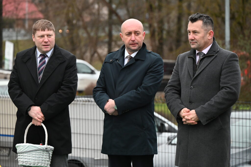 Trzech mężczyzn w ciemnych płaszczach, jeden trzyma biały wiklinowy koszyk, za nimi metalowe ogrodzenie i rozmyte tło z samochodami oraz drzewami.