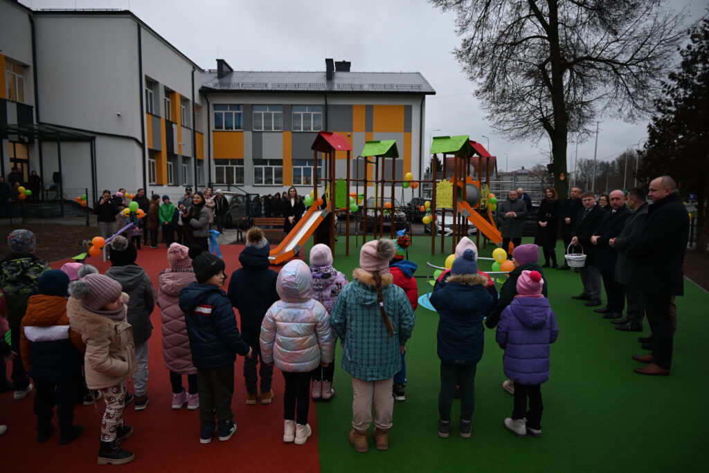 Dzieci w kolorowych kurtkach stojące na placu zabaw z dwoma zjeżdżalniami obok budynku z żółto-szarymi panelami, w tle dorośli i balony.