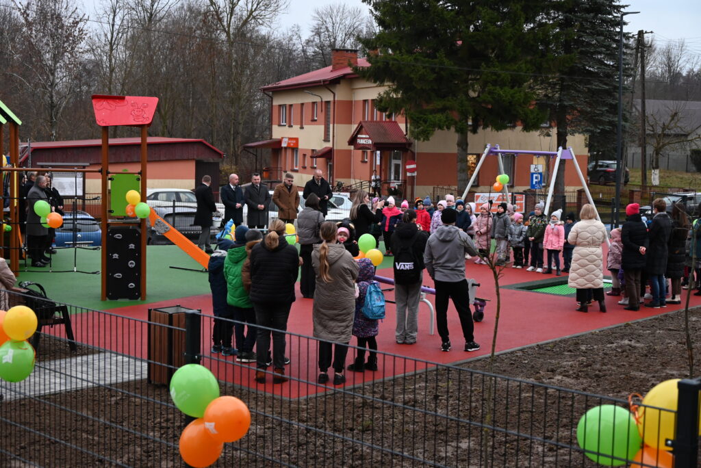 Plac zabaw z kolorową nawierzchnią, huśtawki, zjeżdżalnia, dzieci i dorośli w kurtkach, balony w kolorach zielonym, pomarańczowym i żółtym, budynek w tle.