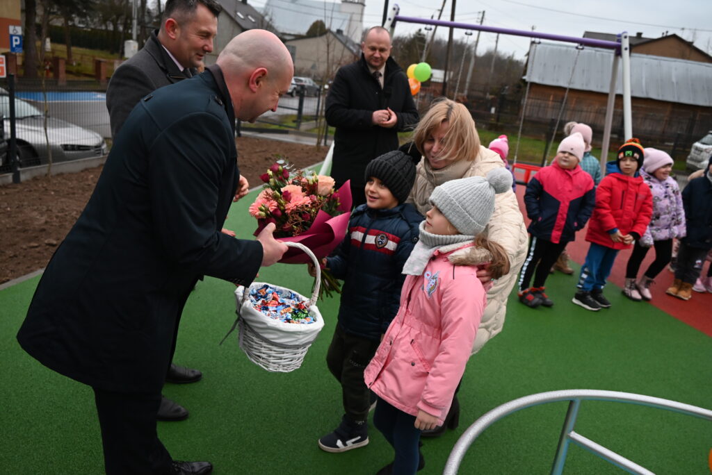 Mężczyzna w czarnym płaszczu podaje dzieciom bukiet kwiatów i koszyk cukierków na zielonej nawierzchni placu zabaw, obok stoją dzieci i kobieta w jasnej kurtce.