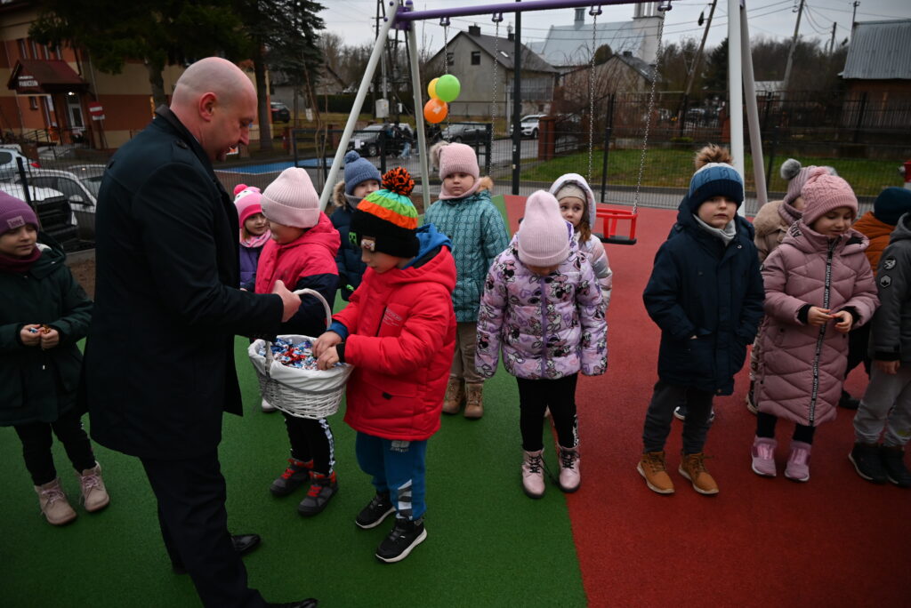 Mężczyzna w czarnym płaszczu rozdaje dzieciom cukierki z białego koszyka na zielono-czerwonej nawierzchni placu zabaw, dzieci w kolorowych kurtkach i czapkach.