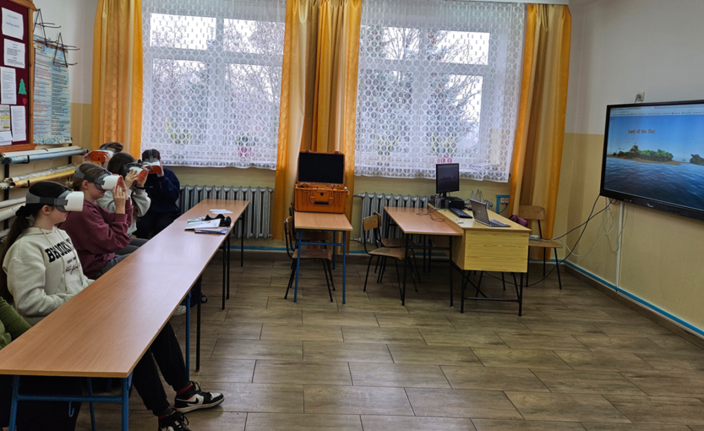 Grupa osób siedząca przy długim drewnianym stole, każda z białymi okularami VR, za nimi żółte zasłony i okna z białą firanką, po prawej ekran z widokiem wyspy i napisem "Lord of the flies".