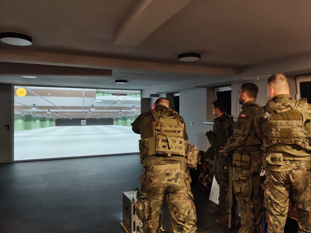 Czterech żołnierzy w mundurach kamuflażowych stoi w pomieszczeniu, jeden celuje w ekran projekcyjny z symulacją strzelnicy.