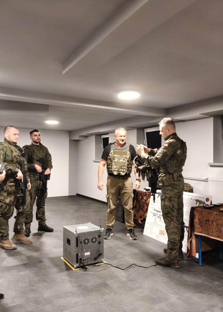 Czterech mężczyzn w mundurach wojskowych i kamizelkach taktycznych w jasnym pomieszczeniu z szarą podłogą, metalowe urządzenie na podłodze z przewodami.