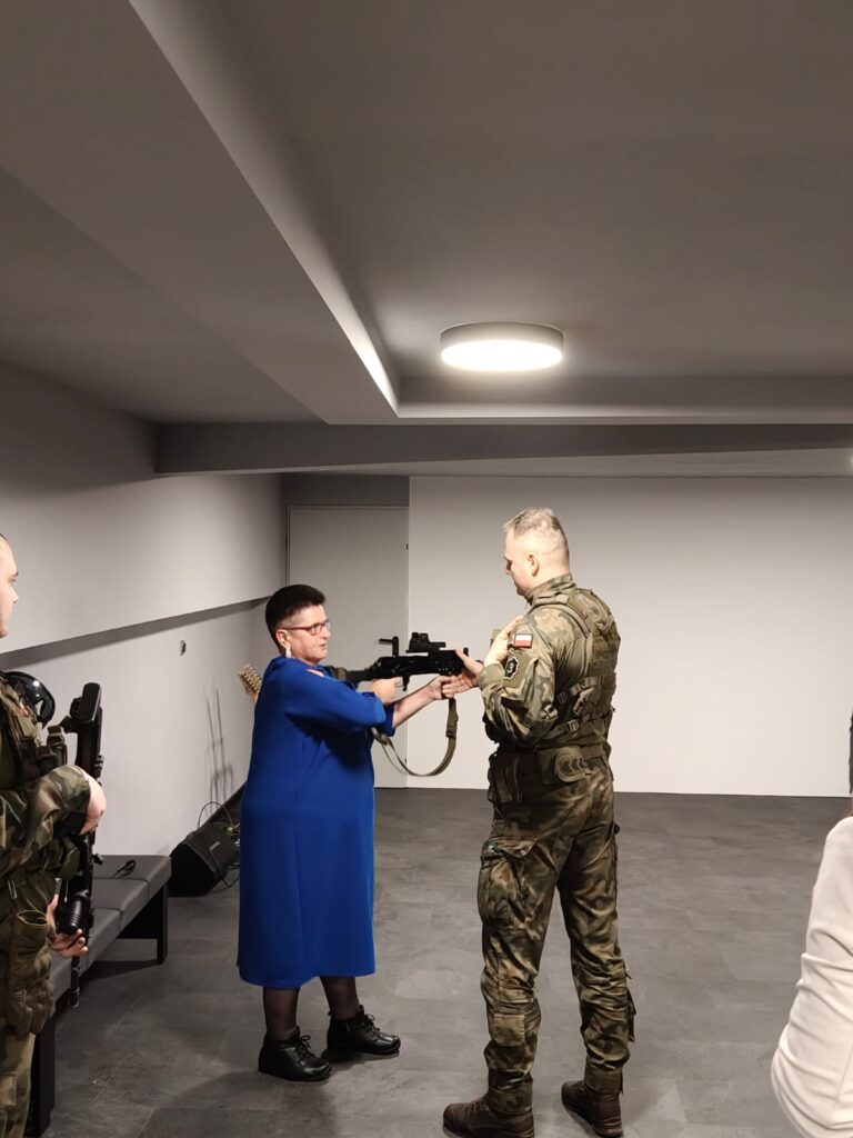 Kobieta w niebieskiej sukience trzyma karabin, stoi naprzeciw żołnierza w mundurze kamuflażowym, w tle jasne ściany i sufit z okrągłą lampą.