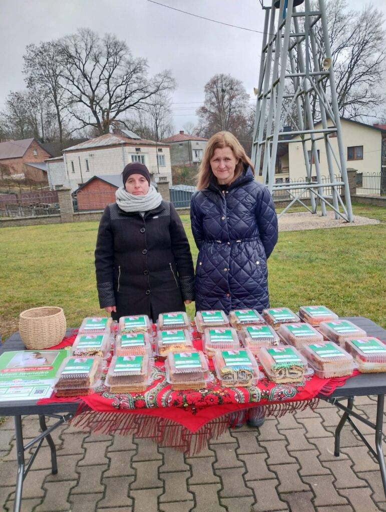 Dwie kobiety w ciemnych kurtkach stoją za stołem z plastikowymi pojemnikami na ciasta, na stole czerwona chusta, w tle domy i metalowa konstrukcja.