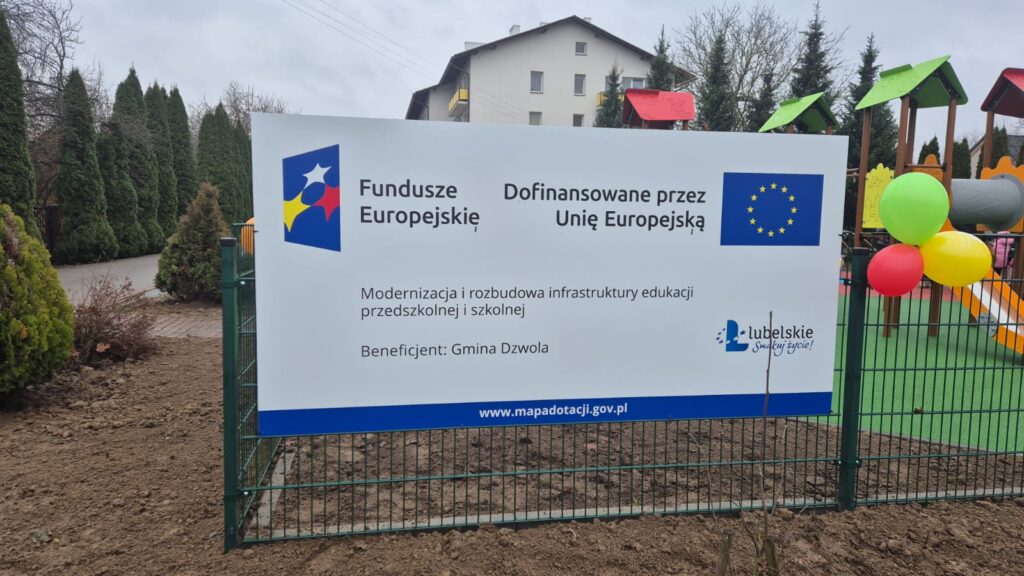 Tablica informacyjna na ogrodzeniu placu zabaw z kolorowymi balonami, z napisem o dofinansowaniu Unii Europejskiej i logotypami, w tle dom i drzewa iglaste.