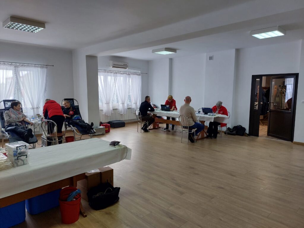 Jasne pomieszczenie z drewnianą podłogą, białe ściany, trzy pary osób, dwie na leżankach, trzy przy stole, białe zasłony, metalowe krzesła, czerwone i czarne torby na podłodze.