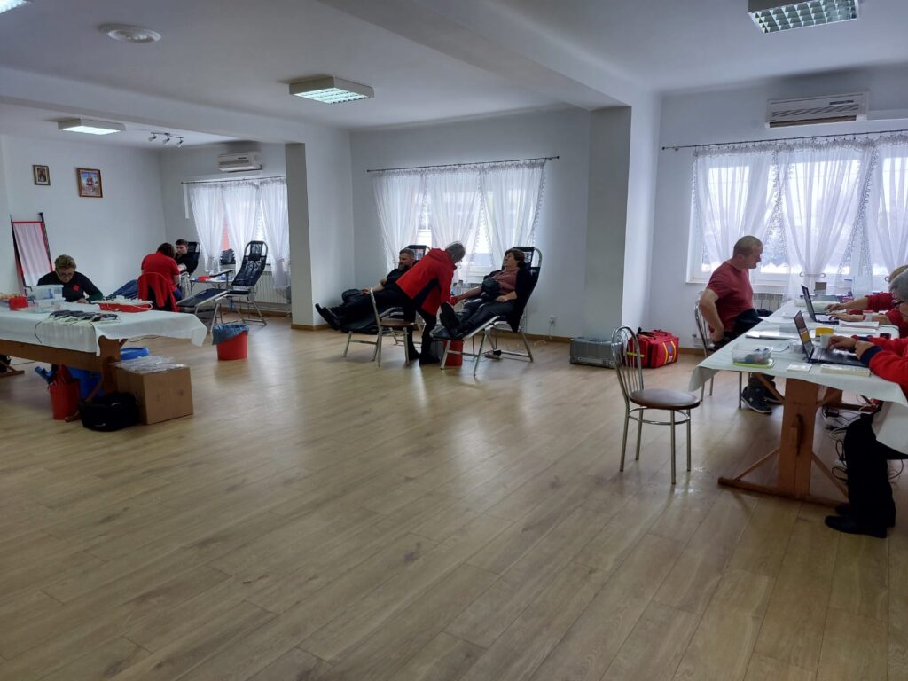 Pomieszczenie z jasnymi ścianami i dużymi oknami z białymi zasłonami, drewniana podłoga, kilka osób na krzesłach medycznych, stoły z laptopami i sprzętem medycznym.