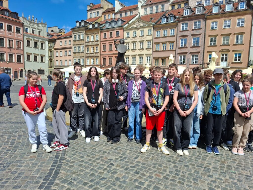 Grupa młodzieży stojąca na brukowanym placu, w tle kolorowe kamienice i pomnik, niebo błękitne z niewielkimi chmurami.