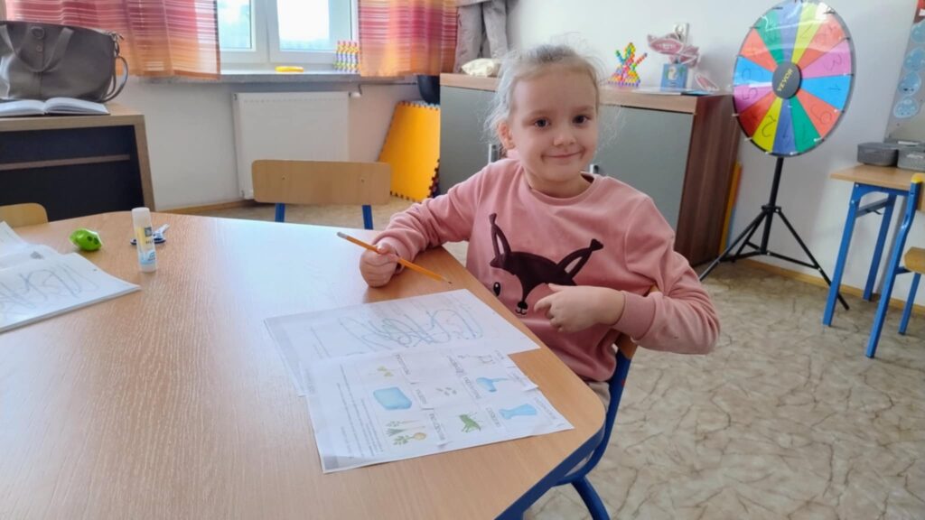 Dziewczynka w różowej bluzie z czarnym nadrukiem siedzi przy drewnianym stole z kartkami i ołówkiem, w tle kolorowe koło fortuny i meble szkolne.