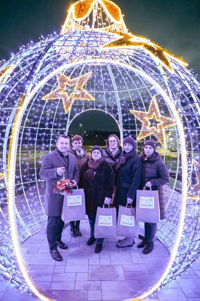 Grupa sześciu osób w zimowych ubraniach stoi w świetlnej dekoracji w kształcie bombki z gwiazdami, trzyma torby z napisem „WESOŁYCH ŚWIĄT”.