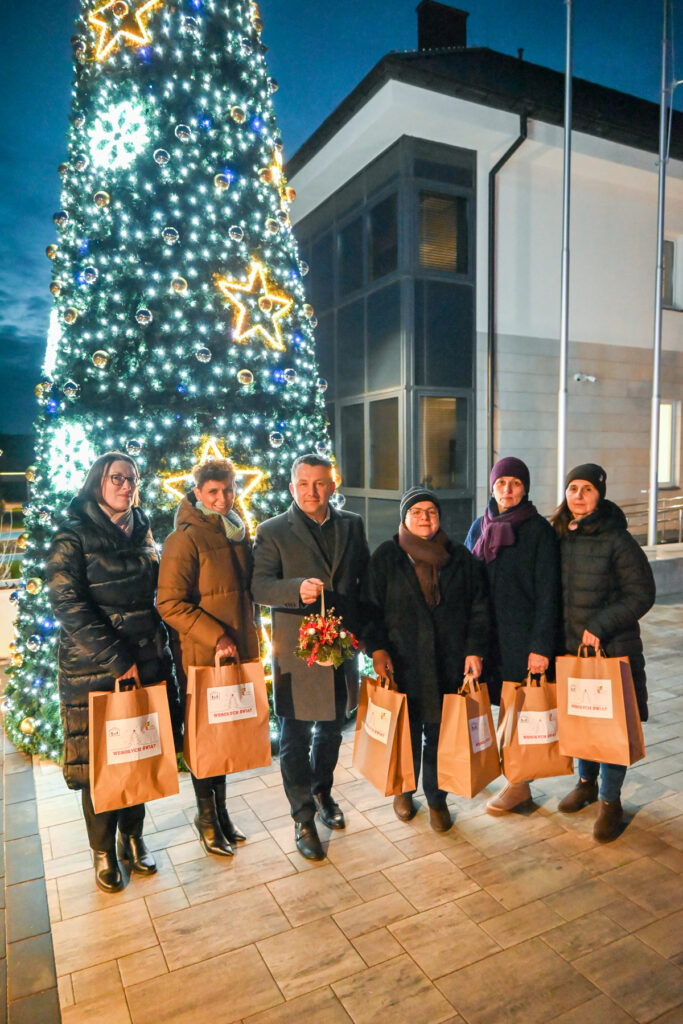Grupa sześciu osób w zimowych ubraniach stoi na tle dużej, udekorowanej i oświetlonej choinki, trzymając papierowe torby z napisem "Wesołych Świąt".