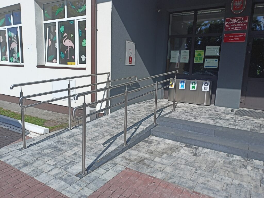 Wejście do budynku z podjazdem dla niepełnosprawnych, szare płytki, metalowe poręcze, po prawej trzy pojemniki do segregacji śmieci, okna z dekoracjami różowego flaminga i zielonych liści.