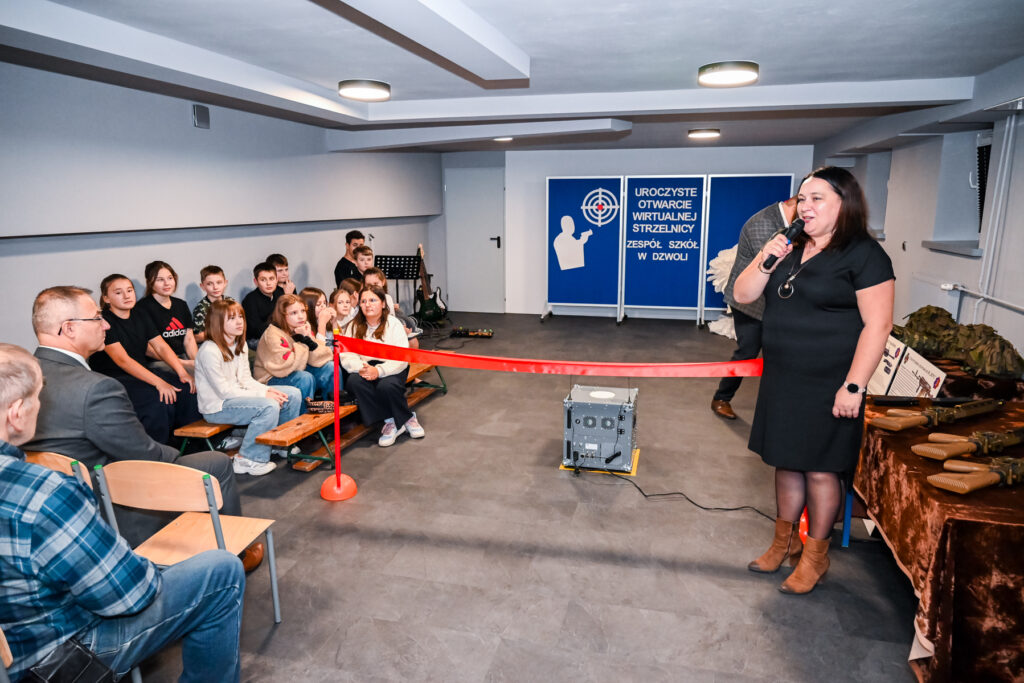 Grupa dzieci i dorosłych siedzących na ławkach w jasnym pomieszczeniu, kobieta z mikrofonem w czarnej sukience stoi obok stołu z atrapami broni, czerwony sznurek rozciągnięty.