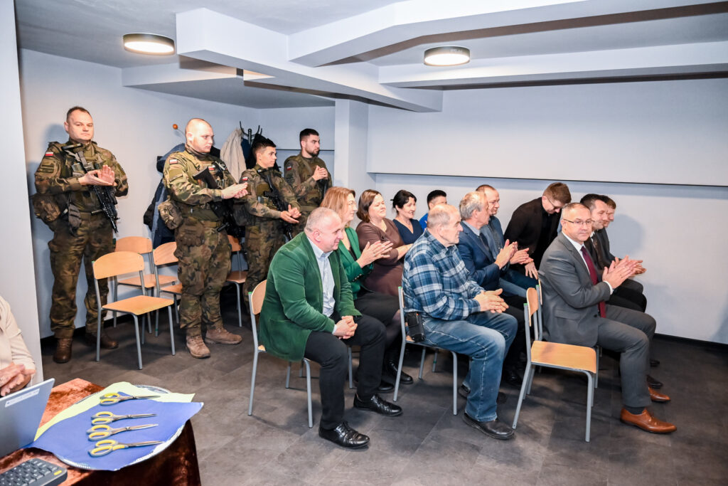 Grupa ludzi siedzi na krzesłach w sali, część osób ubrana w mundury wojskowe stoi z tyłu, na stole leżą narzędzia do cięcia, ściany i sufit białe, podłoga ciemna.