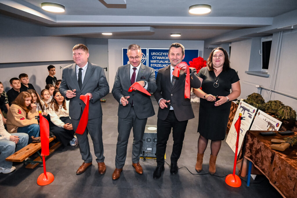 Cztery osoby trzymające nożyczki i czerwone wstęgi, za nimi siedzi grupa dzieci, na stole po prawej hełmy i repliki broni.