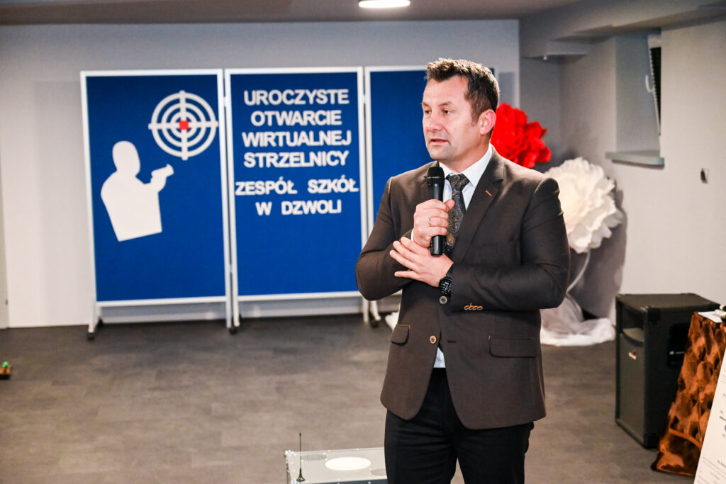 Mężczyzna w marynarce i krawacie mówi do mikrofonu, w tle niebieskie plansze z napisem i białą sylwetką celującej osoby.