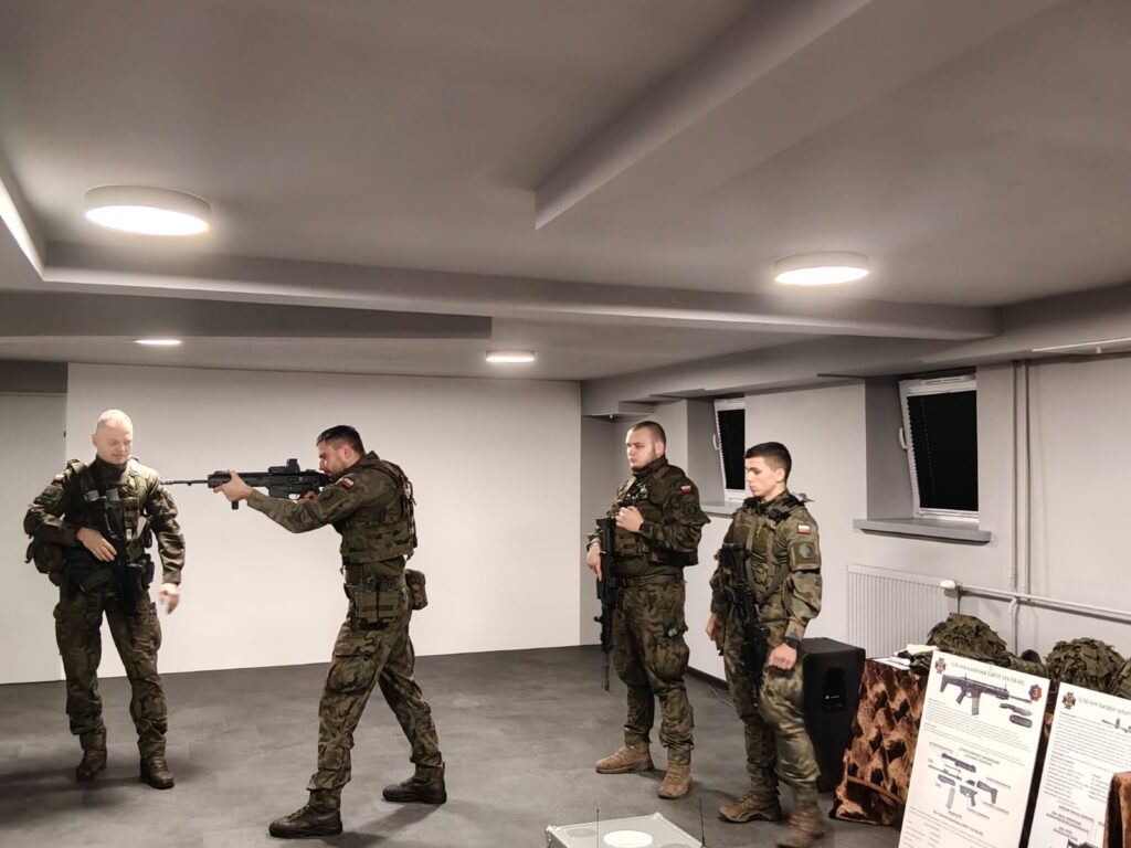 Czterech żołnierzy w mundurach kamuflażowych w jasnym pomieszczeniu, jeden z nich celuje z broni, na stole obok plansze informacyjne i sprzęt wojskowy.