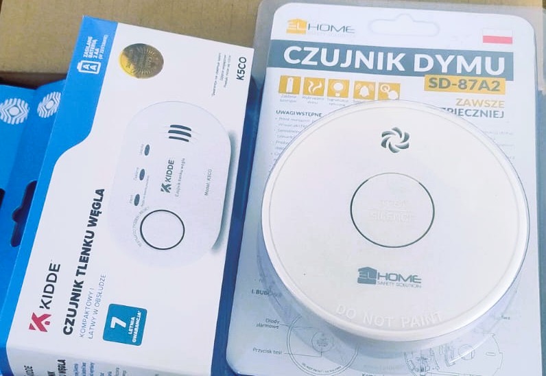 Opakowania białych czujników: czujnik tlenku węgla Kidde i czujnik dymu 2L Home, teksty w kolorach niebieskim, czarnym i żółtym.
