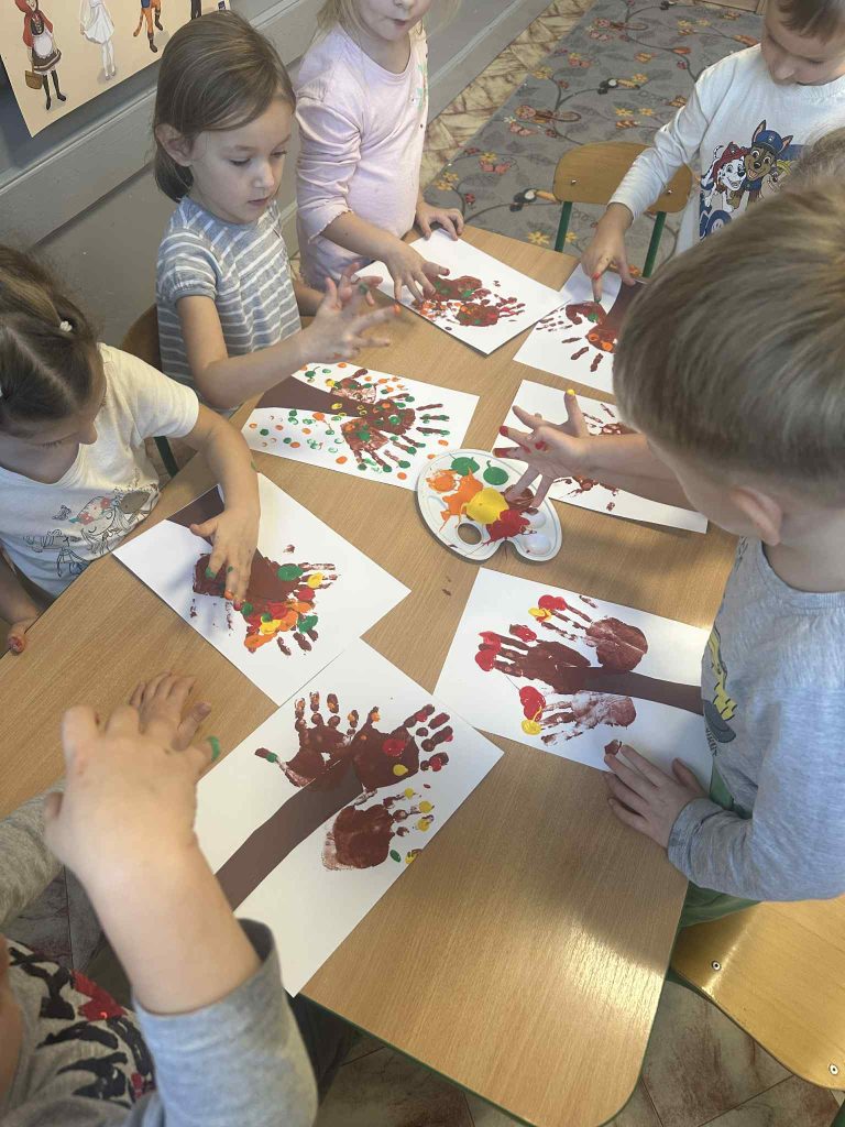 Siedmioro dzieci maluje farbami na białych kartkach, tworząc drzewa z brązowych pni i kolorowych odcisków dłoni na stołach w klasie.