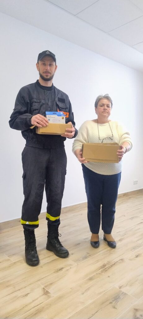 Mężczyzna w czarnym uniformie z żółtymi paskami i kobieta w białym swetrze i granatowych spodniach trzymają kartony na jasnej podłodze w jasnym pomieszczeniu.