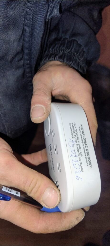 Dłonie trzymające biały czujnik dymu z napisem w języku polskim i podpisaną datą instalacji, ubranie z ciemnego materiału.