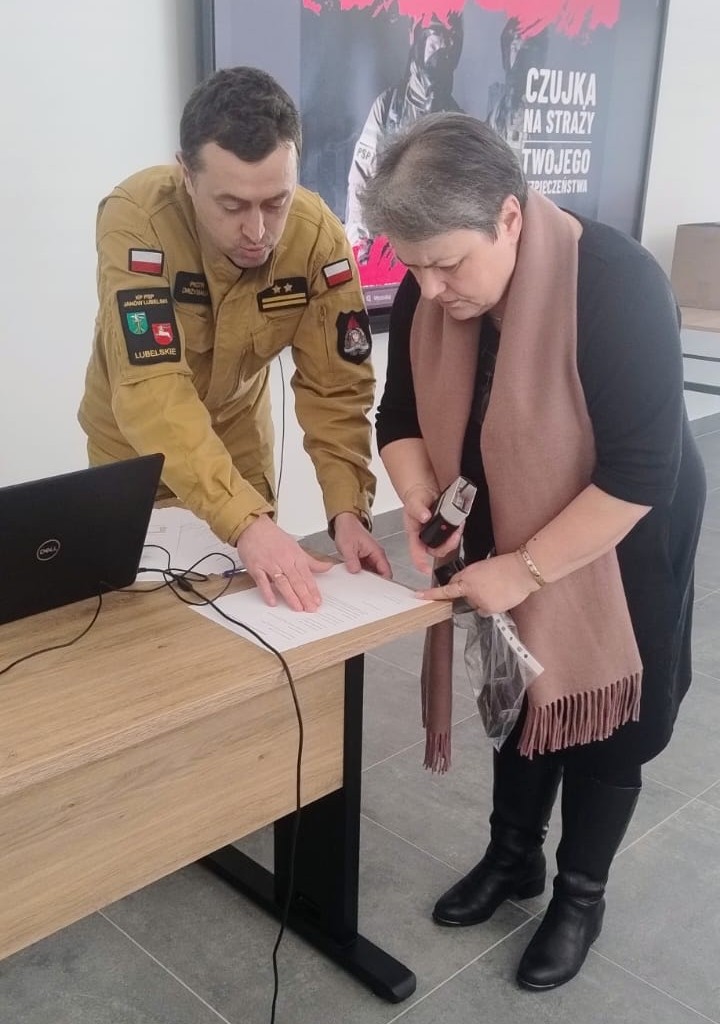 Mężczyzna w mundurze koloru khaki i kobieta w czarnej sukience z różowym szalem pochylają się nad dokumentem na drewnianym biurku, obok stoi czarny laptop.