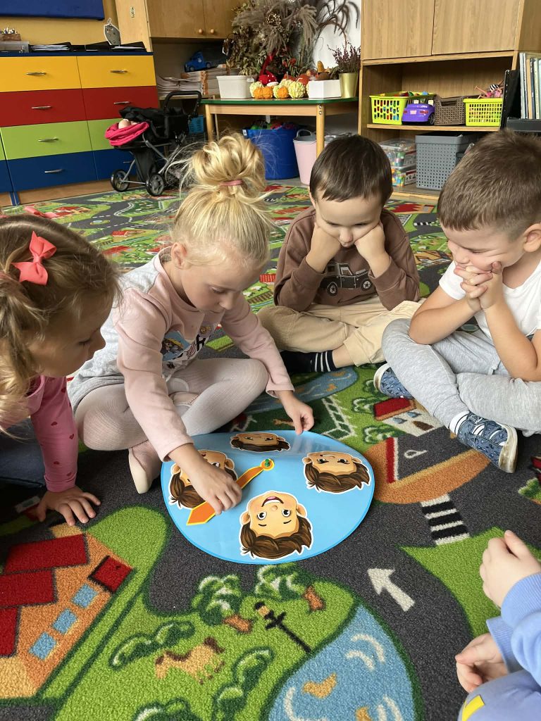 Czworo dzieci siedzi na kolorowym dywanie z motywem miasteczka, skupione na niebieskiej planszy z ilustracjami twarzy i pomarańczową wskazówką.