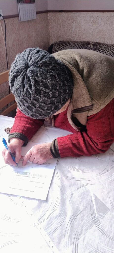 Starsza osoba w czerwonym swetrze, beżowej kamizelce i szarej czapce pisze długopisem na dokumentach na białym obrusie z szarymi wzorami.
