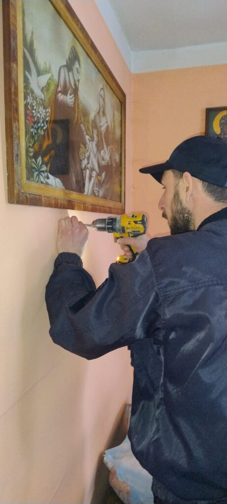 Mężczyzna w czarnej czapce i kurtce używa żółto-czarnej wiertarki do wiercenia w jasnoróżowej ścianie pod obrazem religijnym w drewnianej ramie.