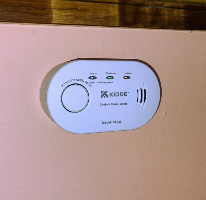 Biały czujnik tlenku węgla KIDDE model K5CO na różowej ścianie, z trzema diodami i okrągłym przyciskiem testu, pod drewnianą ramą.