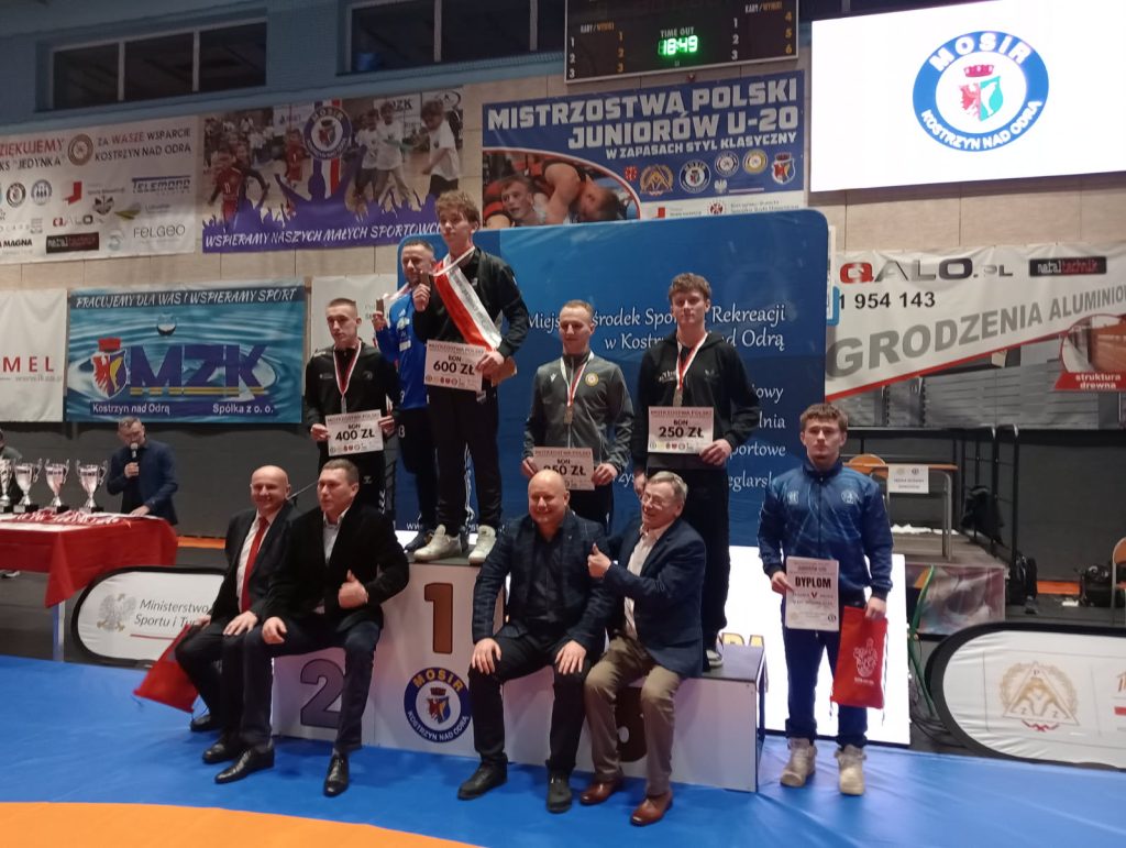 Pięciu młodych mężczyzn na podium z medalami i dyplomami, w tle banery z napisami i logo MOSiR Kostrzyn nad Odrą, kilku mężczyzn siedzi przed podium.