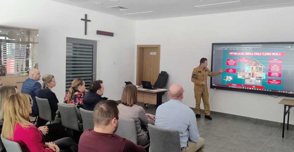 Sala konferencyjna z siedzącymi ludźmi, mężczyzna w mundurze wskazuje ekran z infografiką o źródłach emisji tlenku węgla, jasne ściany, krzyż na ścianie.