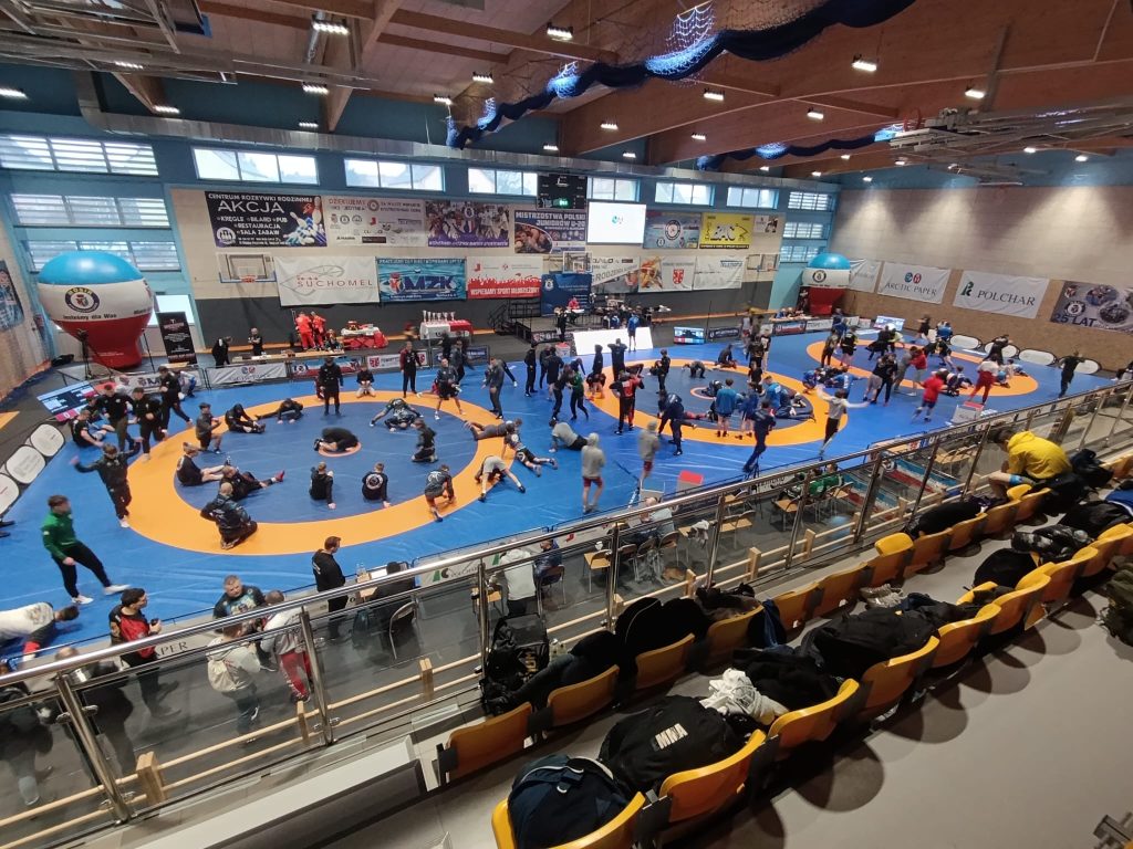 Hala sportowa z niebieskimi i pomarańczowymi matami, wiele osób ćwiczy na matach, widoczne żółte trybuny z porozrzucanymi torbami i ubraniami.