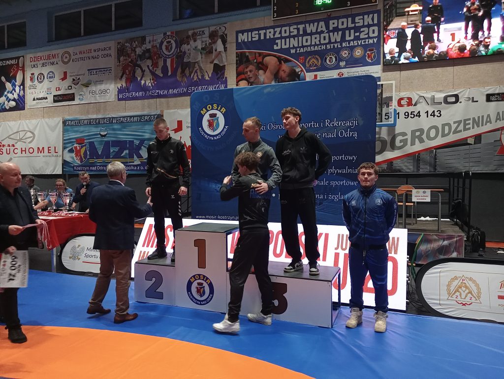 Czterech młodych zawodników na podium w sportowych dresach, w tle banery z napisem Mistrzostwa Polski Juniorów U-20, niebieska i pomarańczowa mata.