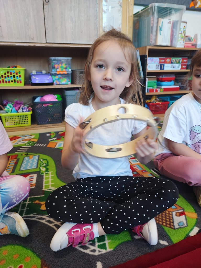 Dziewczynka w białej koszulce i czarnych spodniach w białe kropki siedzi na kolorowym dywanie, trzyma drewnianą tamburynę. W tle półki z pojemnikami i zabawkami.