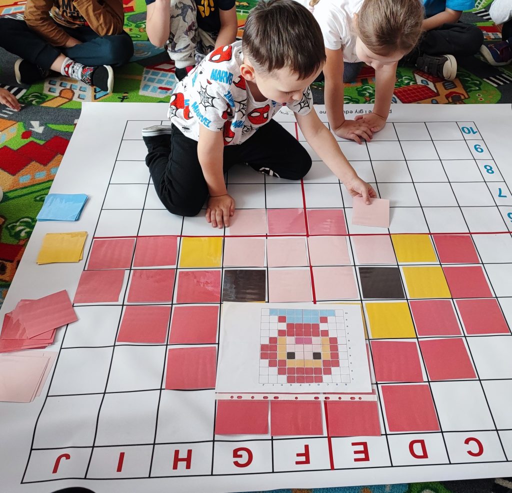 Dzieci układają kwadratowe kolorowe kartki na dużej planszy z oznaczonymi literami i cyframi, tworząc wzór przypominający postać z czerwonymi, żółtymi i czarnymi elementami.