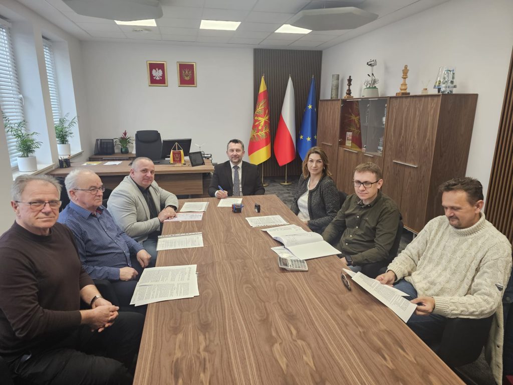 Siedem osób przy długim drewnianym stole w jasnym biurze z trzema flagami i szafą z dokumentami w tle.