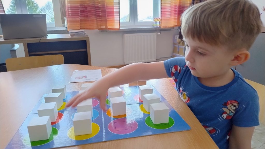 Chłopiec w niebieskiej koszulce układa białe kostki na kolorowej planszy na drewnianym stole w jasnym pokoju z zasłonami w paski.