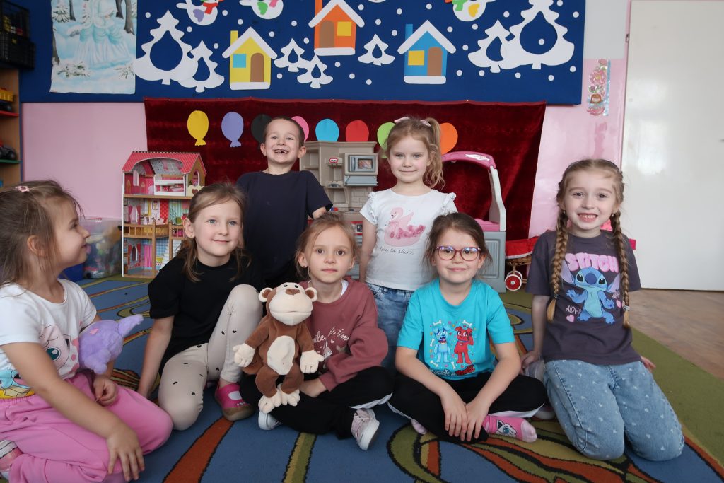 Siedmioro dzieci na kolorowym dywanie w pokoju z zabawkami, na ścianie dekoracje z domkami i choinkami, dwie dziewczynki trzymają pluszaki.