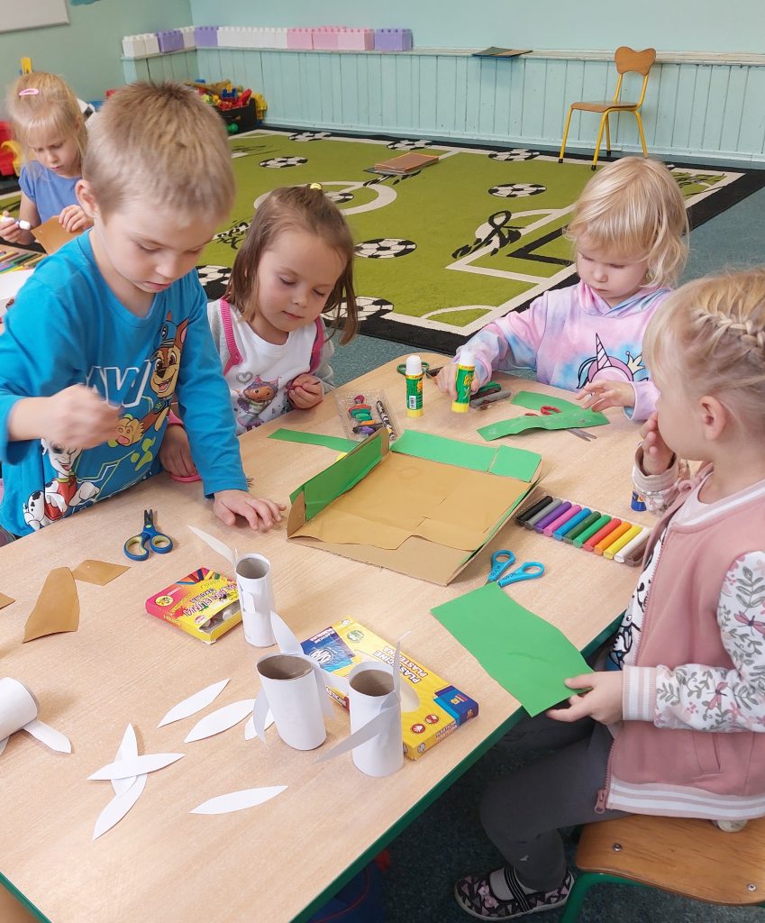 Cztery dzieci siedzące przy drewnianym stole z nożyczkami, klejem, kolorowym papierem i plasteliną, w tle zielony dywan z motywem piłek nożnych.